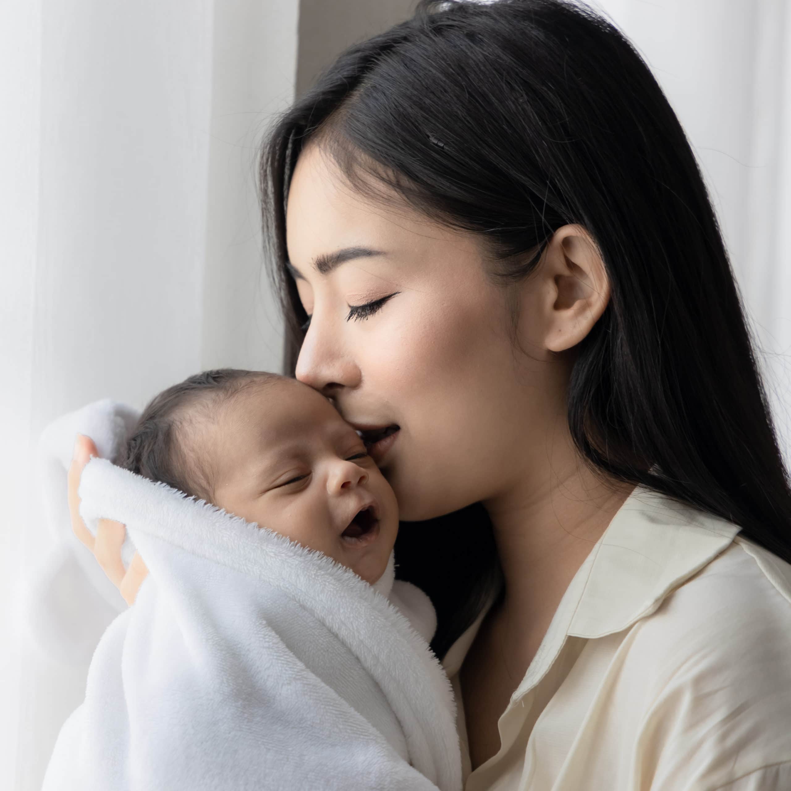 Portrait of asian young mother kissing her cute newborn baby in white bed room. Wife and mother's day concept.