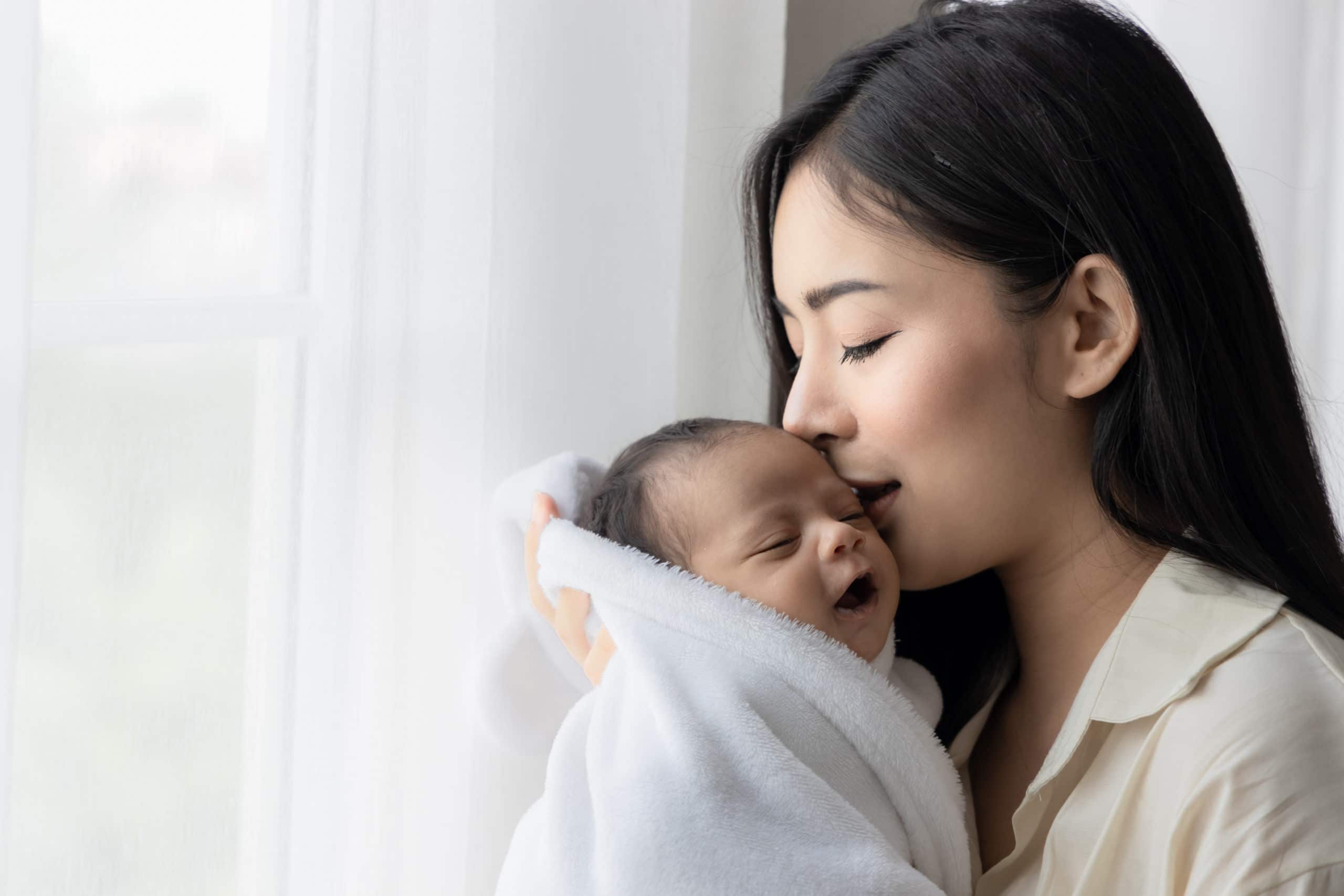 Portrait of asian young mother kissing her cute newborn baby in white bed room. Wife and mother's day concept.