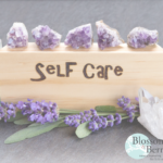 Picture of a piece of wood with the word self care on surrounded by crystals and flowers